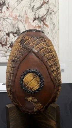 Orythi: Dragon's Egg by Tzontli, an oval brown sculptural object with layered carved textures on a dark wood stand.