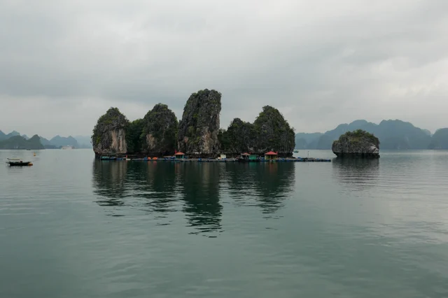 Untitled photograph by Mariana De la Vega — limestone karst islands rising from still, misty water, small floating boats gathered at their base.