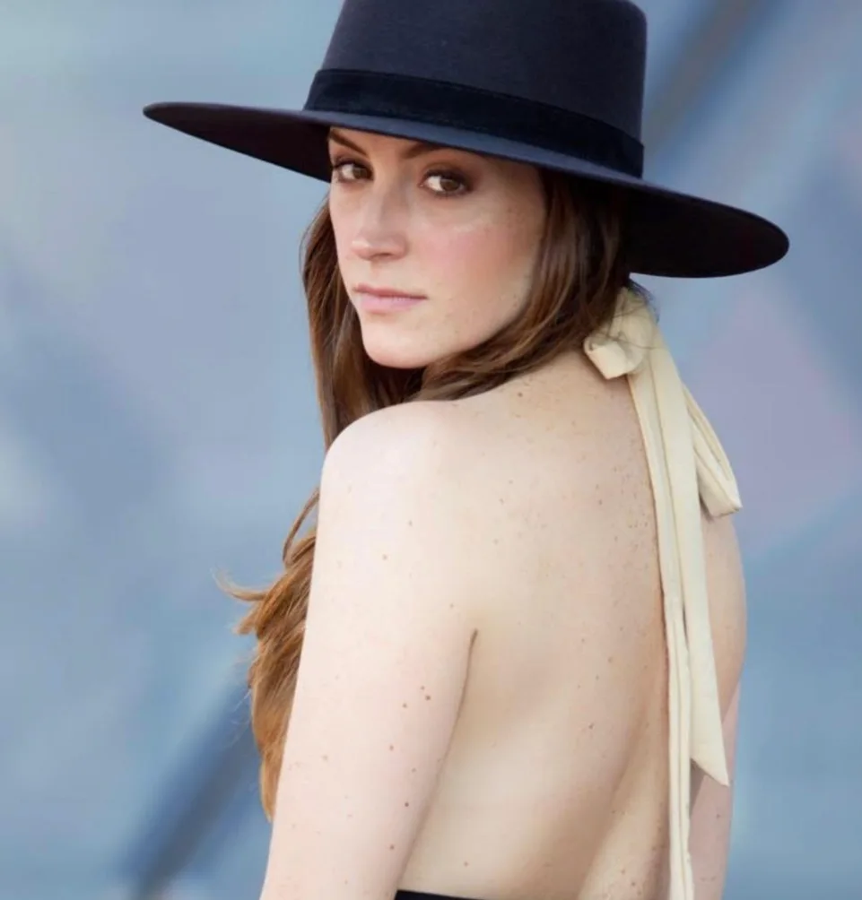 Portrait of Mariana De la Vega wearing a wide-brimmed black hat, looking over her shoulder against a soft blue sky.