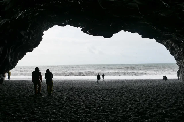 Untitled photograph by Mariana De la Vega — figures silhouetted at the mouth of a sea cave, looking out across a dark volcanic shore to breaking ocean waves.