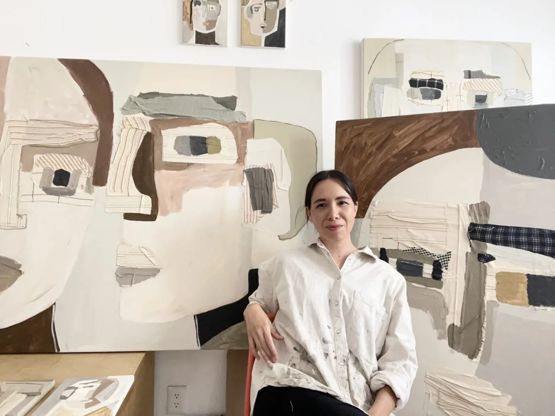 Portrait of Raquel Barrios seated in her studio in front of large mixed-media abstract paintings.